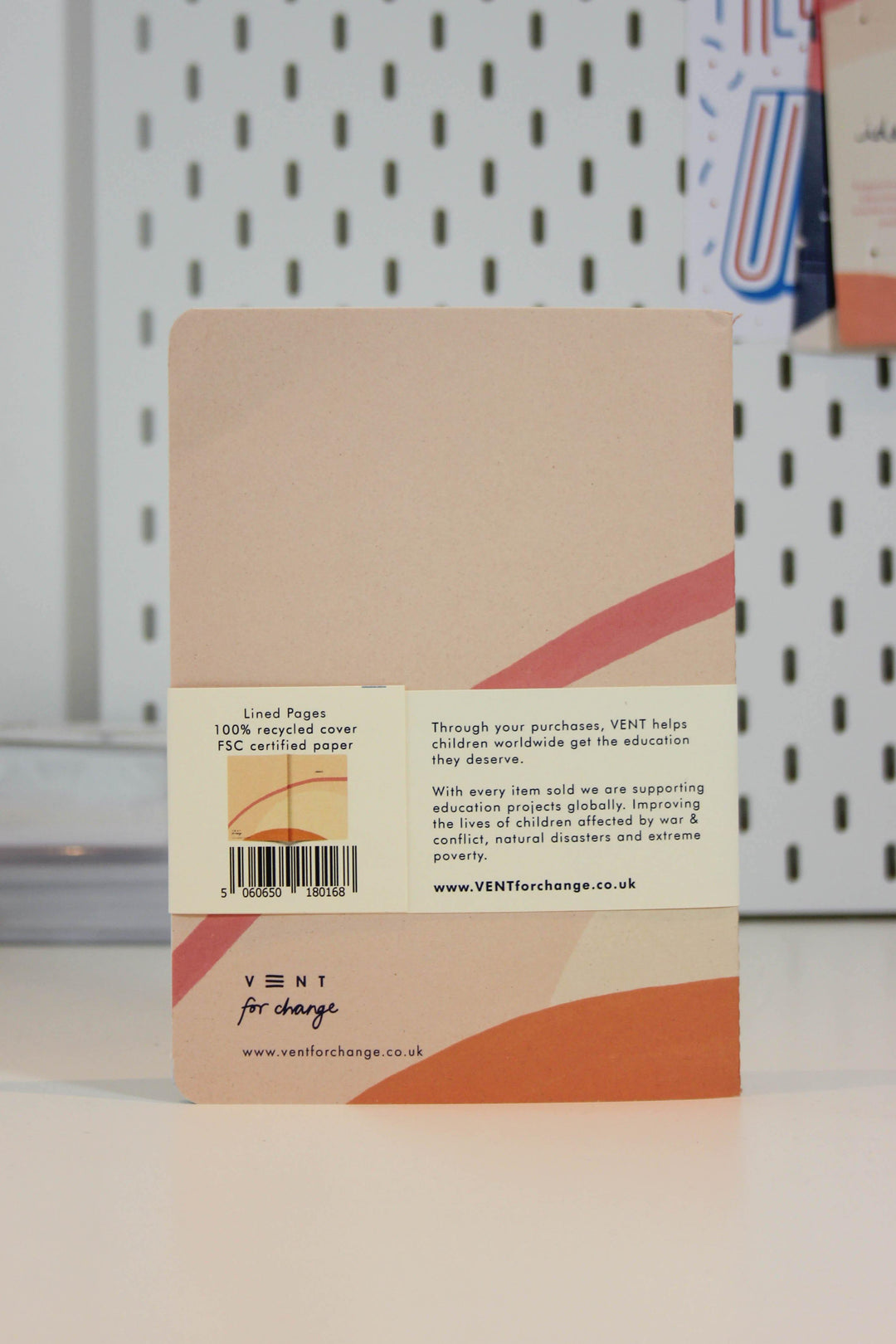 A Recycled Soft Cover A5 Notebook – Ideas Cream from Vent For Change with a cardboard cover and red-orange diagonal stripes sits on a white surface against a pegboard background. A label on the back details the notebook as having lined pages and an FSC certified recycled cover, mentioning VENT's support for children's education globally.