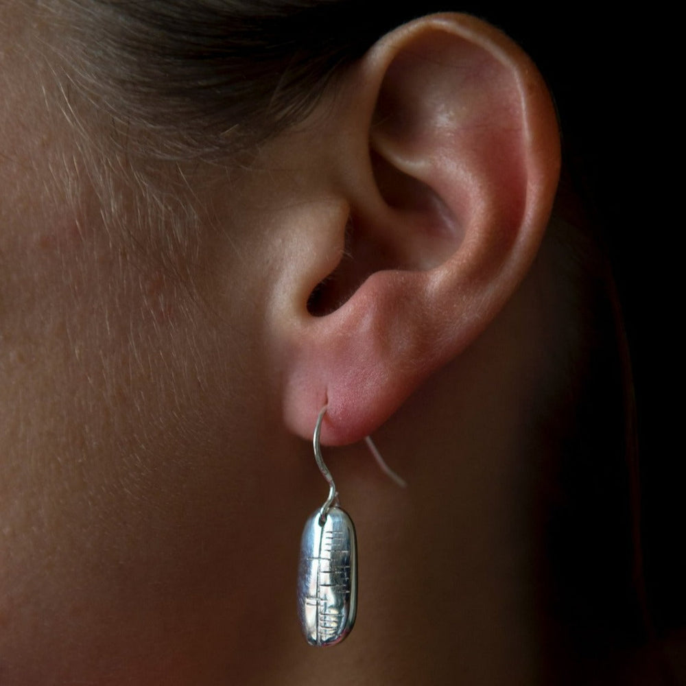 Close-up of a person's ear wearing a dangling sterling silver earring. The Ogham Treasure Happiness (Sonas) Carved Earring features an elongated oval shape with a textured, reflective surface that catches the light. The background is dark, highlighting the earring and ear. Perfectly gift boxed for any occasion.