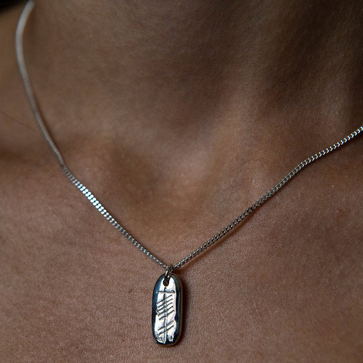 Close-up of a person wearing a sterling silver necklace with a rectangular Love (Grá) Carved Silver Pendant from Ogham Treasure. The pendant features a delicate engraved design of the Gaelic word for love. The background shows the person's neck and part of their collarbone, highlighting the simplicity and elegance of the jewelry.