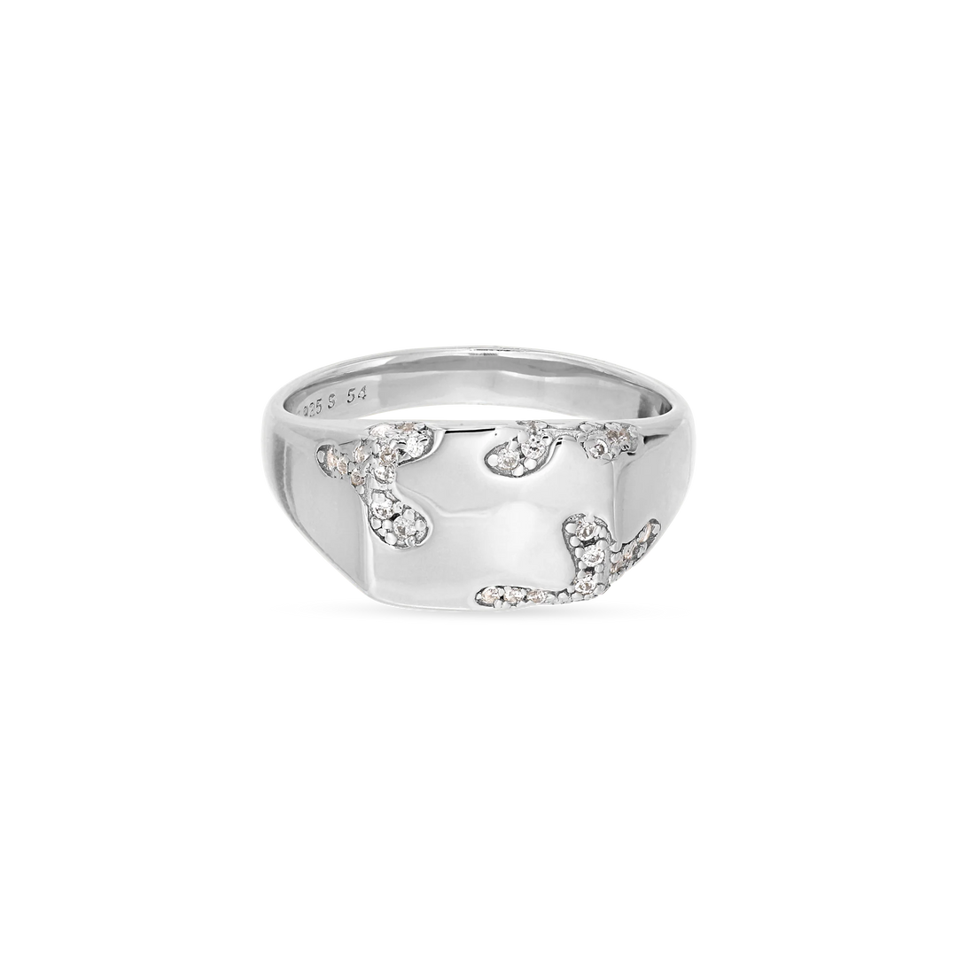 The Iced Signet Ring by Jane Koenig is crafted from 925 sterling silver and features a wide, shiny band adorned with an abstract pattern of small, sparkling white stones.