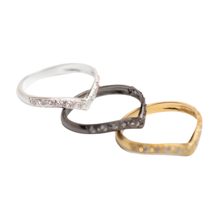 Three Imagination Wishbone Rings with zircon in silver, black, and gold tones by Georgia Charal overlap on a white background, each featuring chevron shapes adorned with sparkling embedded gemstones.
