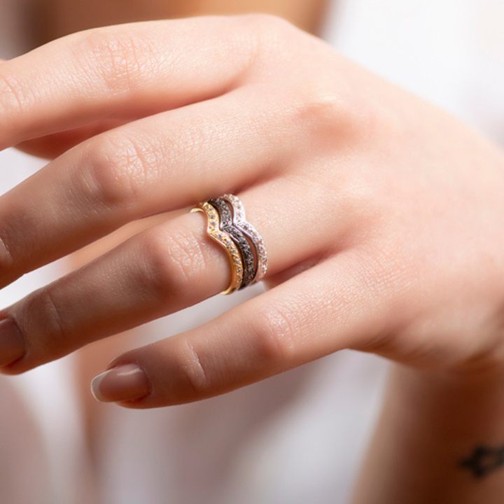 A close-up of a hand wearing two stacked, wavy Georgia Charal Imagination Wishbone Rings with colorful zircon stones in sterling silver, resting against a soft, blurred background.