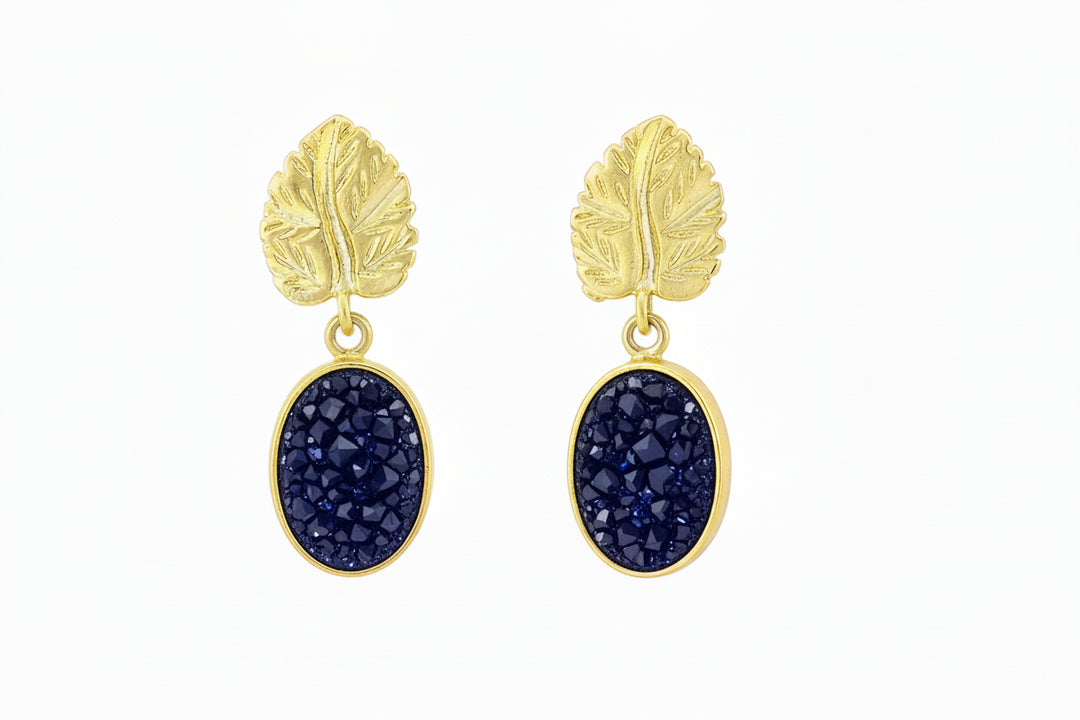 Watermelon tropical Indigo Blue Druzy And Gold Leaf Earrings feature oval indigo druzy stones set in gold plated brass with textured, leaf-shaped tops. Shown on a white background.