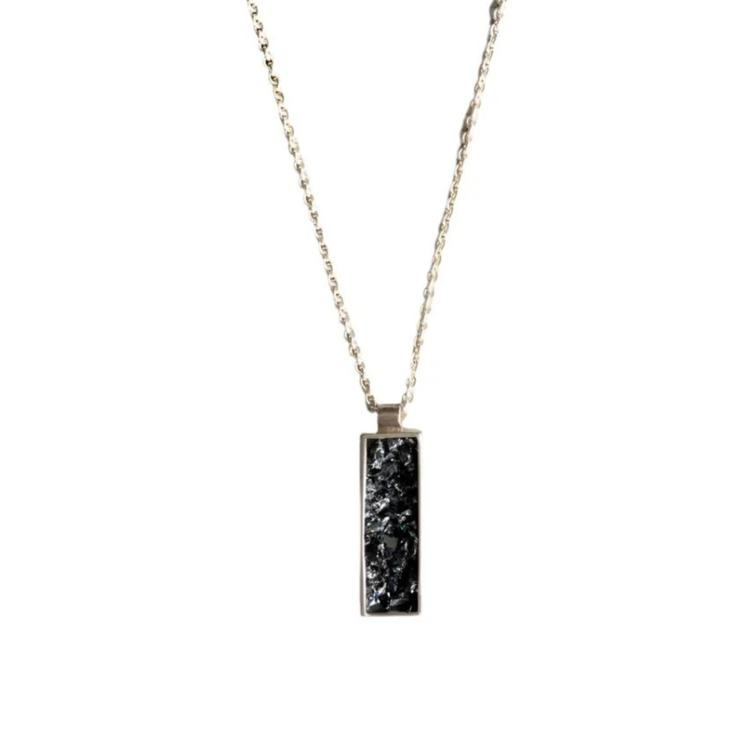 An Ingot Pendant by Angela O'Keefe is shown. The pendant features a rough black stone, possibly crushed silicon carbide, encased in an intricate silver frame. The necklace chain is visible, set against a background composed of contrasting black and light gray areas, evoking a gothic fairy tale aesthetic.