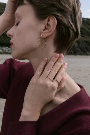 A person with short hair stands outdoors in a burgundy top, showcasing Inner Island's Sister Hoops and multiple rings—ideal for jewellery lovers—with a hand touching their neck and head turned to the side.