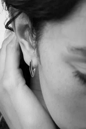 A close-up black and white photo highlights a person's ear adorned with Inner island's Treo Mini Earrings. Their hand gently rests on their neck, eye closed, as soft lighting creates a serene, minimalist mood.
