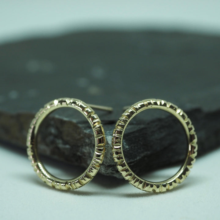 A pair of Aud Into The Light Circle Earrings 9ct Yellow Gold with a textured, spiked design rests against a dark, round rock backdrop. The hand-engraved earrings are circular with intricate, even patterns running along their outer edges, adding a unique and stylish touch.