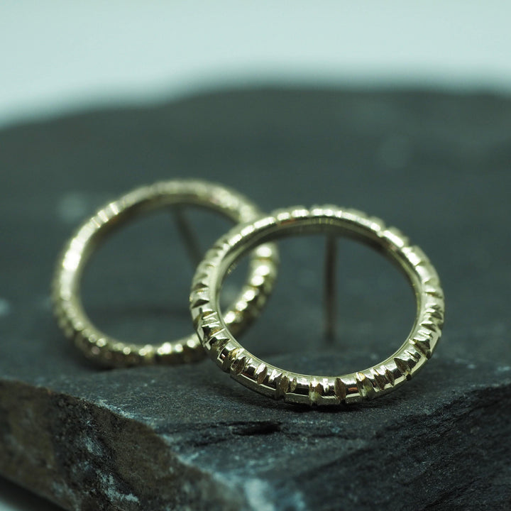 A pair of Into The Light Circle Earrings 9ct Yellow Gold by Aud are displayed on a dark, rocky surface. Hand engraved with an intricate design and a shiny finish, the small earrings stand out against the blurred background, keeping the focus solely on their exquisite craftsmanship.