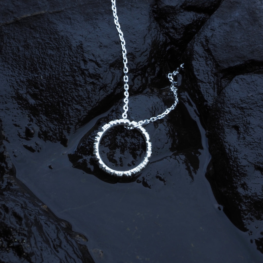 An Into The Light Circle Pendant Silver from Aud with a thin sterling silver chain lies on a textured black rock surface, partially submerged in a small pool of water, reflecting light. The scene creates a minimalistic and elegant aesthetic, highlighting the unique jewelry's craftsmanship.