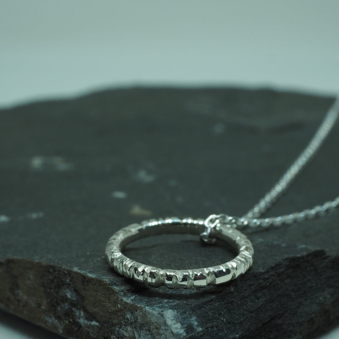 Close-up of a textured sterling silver Into The Light Circle Pendant Silver by Aud on a delicate silver chain, displayed on a dark, rough-surfaced stone. The pendant has a hammered finish, giving it a rustic appearance. This unique jewelry piece stands out as the background is blurred, emphasizing the handmade pendant's intricate details.