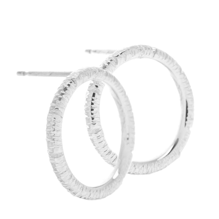 A close-up image of Aud's Into The Light Circle Earrings Silver. The hoops feature a textured, hammered finish, giving them a rustic appearance. The earrings are designed with a simple, circular shape and are positioned overlapping each other in the image.