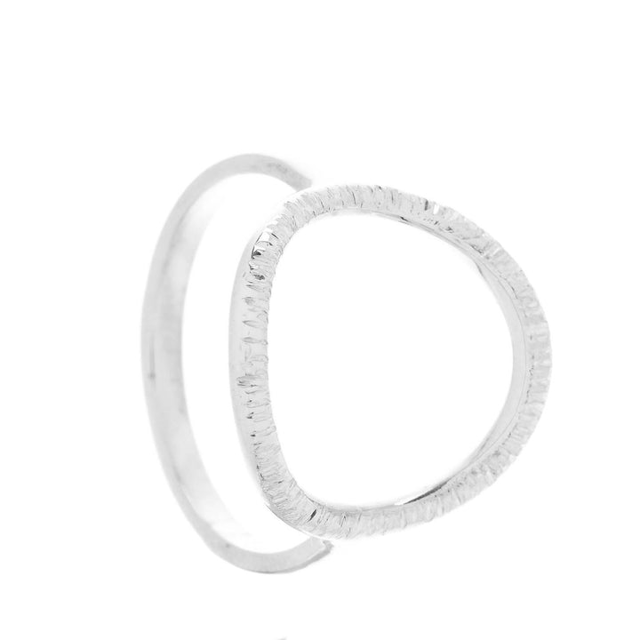 The Aud Into The Light Ring Silver is a handmade sterling silver ring featuring a unique, open geometric design with a textured surface. It has two interconnected loops, creating a modern and minimalist look against a white background.