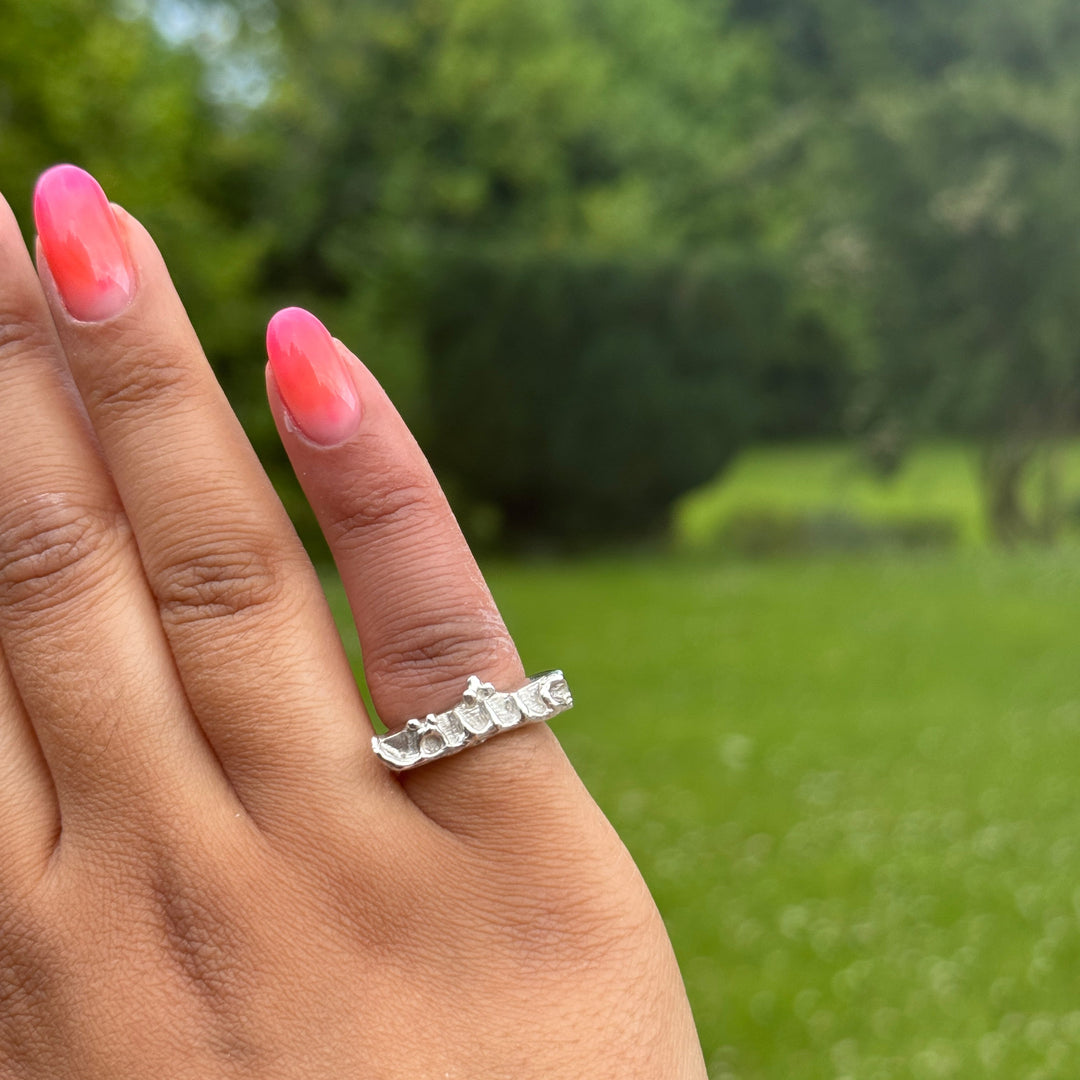 A hand with bright pink nails wears the Ishq Ring by Soufana Attia—a silver twig-shaped piece symbolizing enduring bonds—set against a blurred green outdoor background.