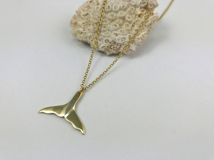 Elegantly displayed on coral, the Jennifer Kinnear Whales Tail Necklace shines with its gold pendant.