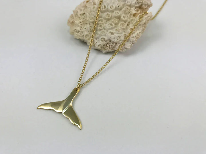 The Jennifer Kinnear Whales Tail Necklace, crafted in gold, is elegantly displayed against coral on a pristine white backdrop.