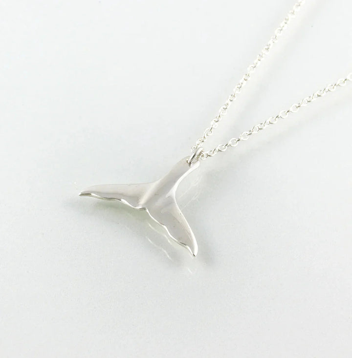 The Jennifer Kinnear Whales Tail Necklace enchants with its silver pendant reflecting light on a smooth surface, displayed on a light background. The delicate chain connects seamlessly to the whale tail fin, embodying Tarifa's breezy charm.