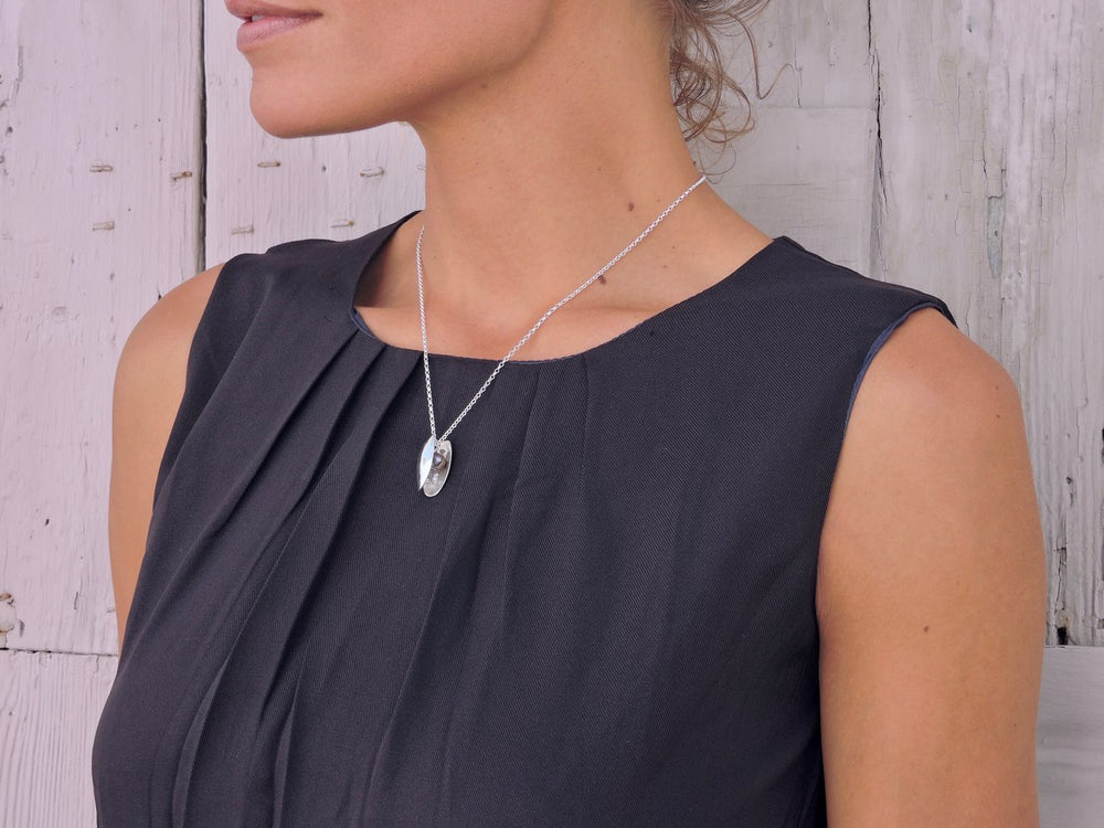 A person with light skin and blonde hair is shown from the neck down, wearing a sleeveless, dark grey dress. Around their neck, they have a stunning Jennifer Kinnear Open Clam Shell Necklace With Pearl in silver. The background is a light-colored, rustic wooden wall.