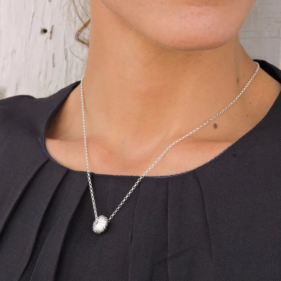 A woman wearing a Jennifer Kinnear Sea Urchin Necklace Small. She is dressed in a dark, pleated top, visible from the shoulders up. The background is a neutral-colored wall with some visible texture.