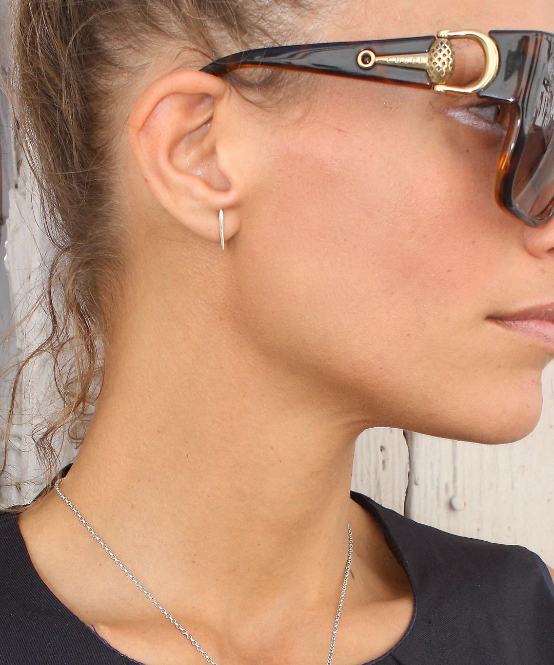 Close-up of a person with light skin wearing oversized tortoiseshell sunglasses with gold accents, Sea Urchin Spine Earrings - Silver by Jennifer Kinnear, and dark clothing. Their hair is pulled back, and they are standing against a neutral, textured background.