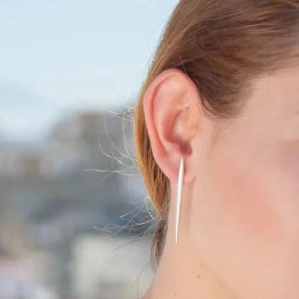 Close-up of a person with light brown hair wearing minimalist, sterling silver earrings. The Sea Urchin Spine Earrings - Silver by Jennifer Kinnear feature a simple straight-line design, going through the earlobe and hanging down. The background is blurred, with a pale blue sky and some indistinct objects.