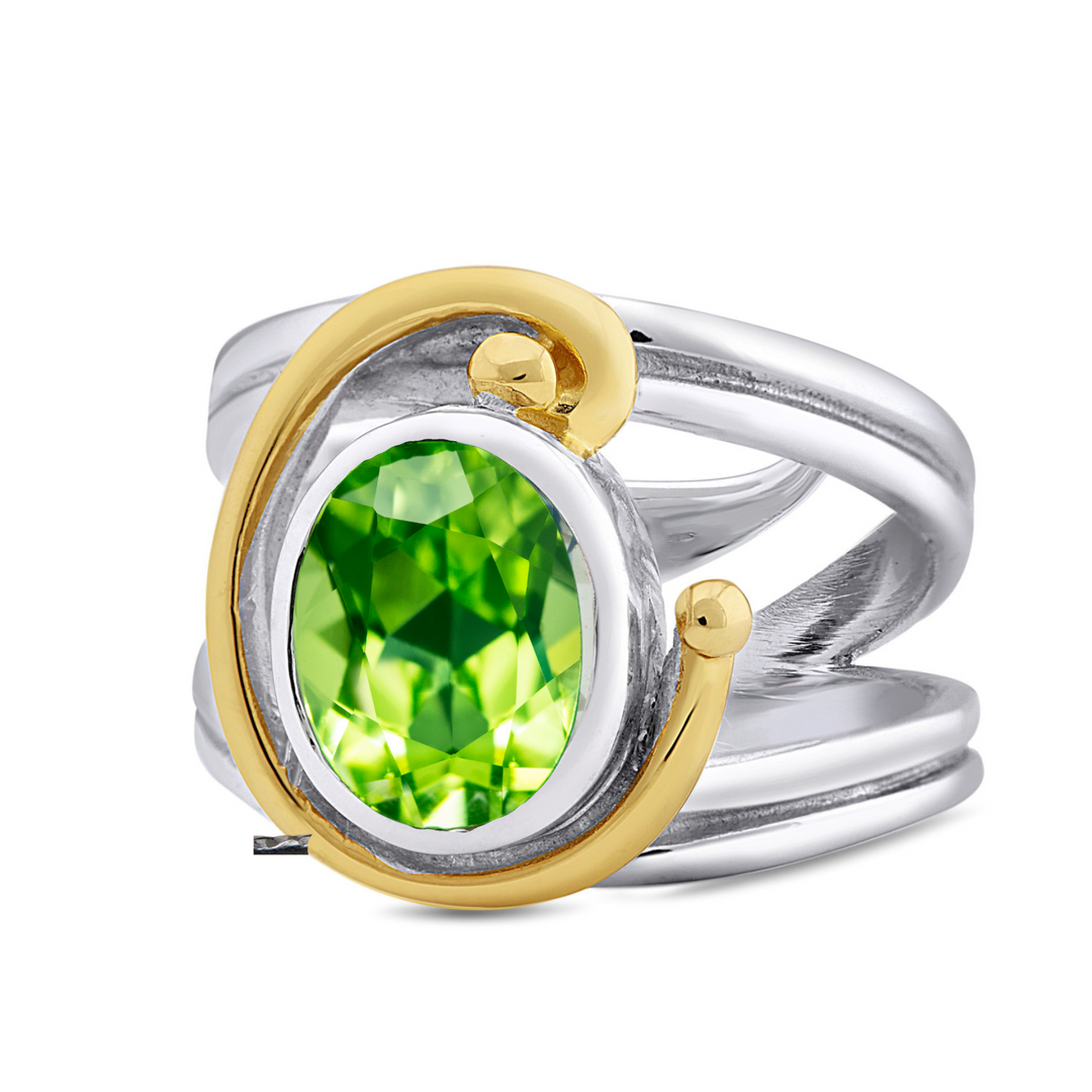 The Adjustable Jasmine Ring by Gallardo & Blaine Designs features an oval green gemstone set on a wide silver band. Embracing Art Nouveau style, it boasts elegant gold swirl designs on the sides, adding charm to this exquisite piece.