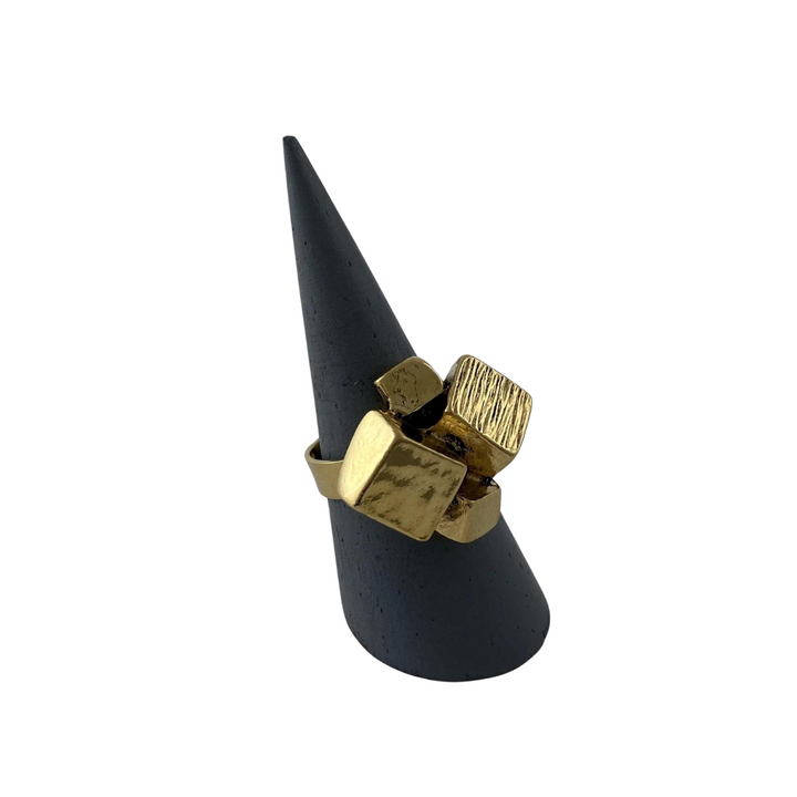 A gold-toned, adjustable Jazz Gold Plated Pewter Ring by J. Boetsch Creations featuring multiple square-shaped, textured elements is displayed on a black, cone-shaped ring holder against a white background.