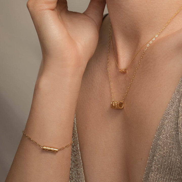 Close-up of a person wearing delicate gold jewelry, including a bracelet with a cylindrical charm, an Andrea Mears Jewellery Karma Wheel Necklace Silver with a small circular charm, and another necklace with a cylindrical charm, all set against a neutral background and glittery fabric.