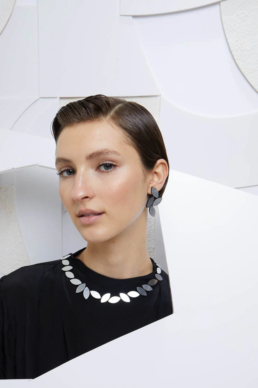 A person with short, slicked-back hair wearing a black top is shown against a geometric white background. They are accessorized with Iskin Sisters' Kate Leaves Earrings Medium, featuring lightweight and comfortable statement earrings with modern, leaf-like shapes. The overall look is sleek and contemporary.