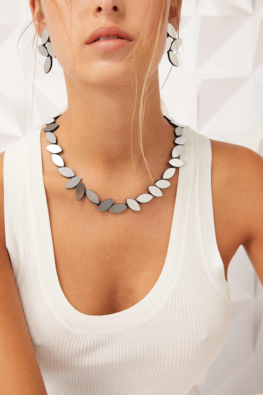 Close-up of a person wearing a sleeveless white top, the Iskin Sisters' Kate Leaves Silver Necklace, and matching earrings with petal-shaped, black and white segments. The background features a geometric white pattern. The person's hair is styled with loose strands falling forward.