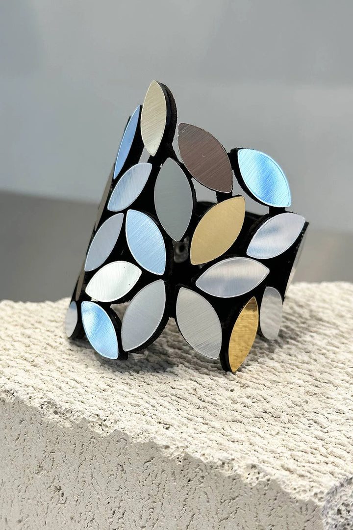 The Kate Leaves Bracelet Medium by Iskin Sisters is a stunning statement piece featuring a pattern of overlapping leaf-shaped pieces in various metallic colors, including silver, gold, and bronze. Presented on a textured stone surface, this bracelet boasts an open-ended design perfect for any occasion.