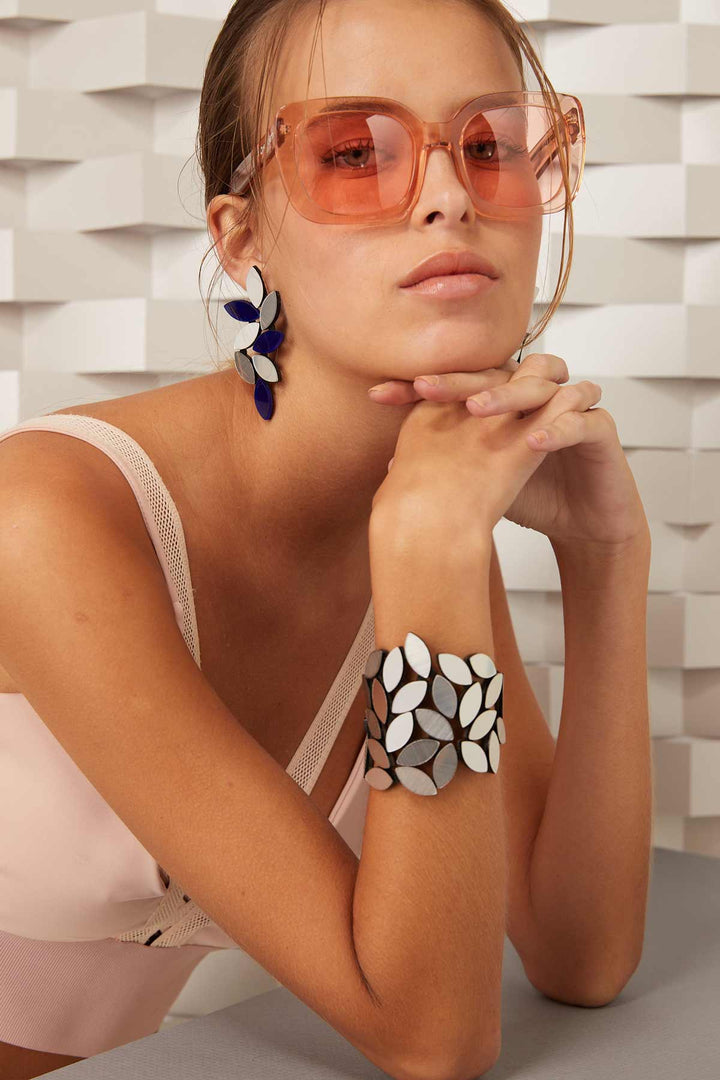 A person with light brown hair is wearing large, translucent, peach-colored sunglasses and resting their chin on their hands. They are also wearing a sleeveless top, large leaf-shaped earrings, and the Kate Leaves Bracelet Medium from Iskin Sisters. The background features a geometric, white pattern.