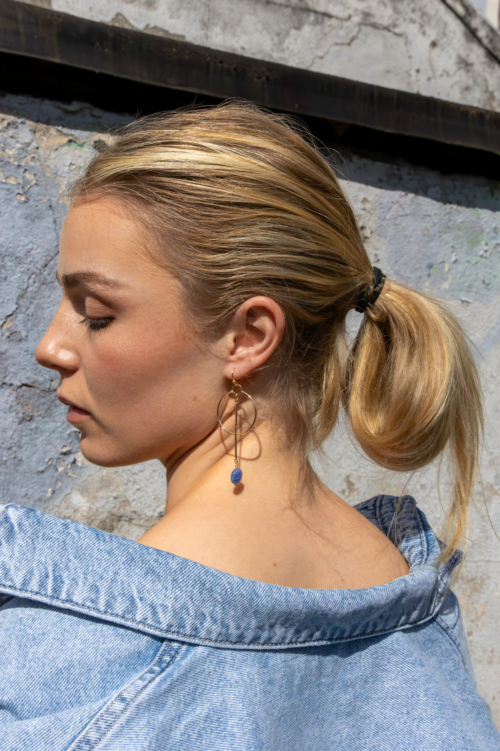 A woman with blonde hair in a ponytail, wearing a blue denim jacket and the Kea Earrings with Sodalite Drop by Kybalion Jewellery, stands sideways before a textured wall with her eyes closed.