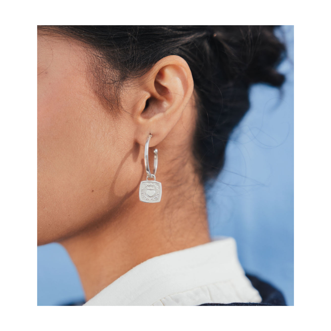 A close-up of a person's ear wearing a silver hoop earring with a square pendant featuring an engraved crest, set against a blue background.