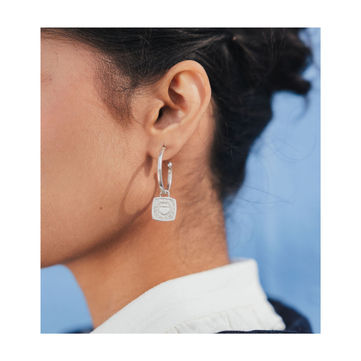 A close-up of a person's ear wearing a silver hoop earring with a square pendant featuring an engraved crest, set against a blue background.