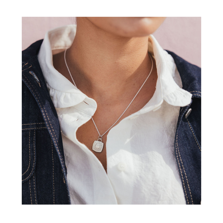 A close-up of a person wearing a delicate silver pendant necklace with a square engraved charm, styled over a white ruffled shirt and a dark denim jacket.