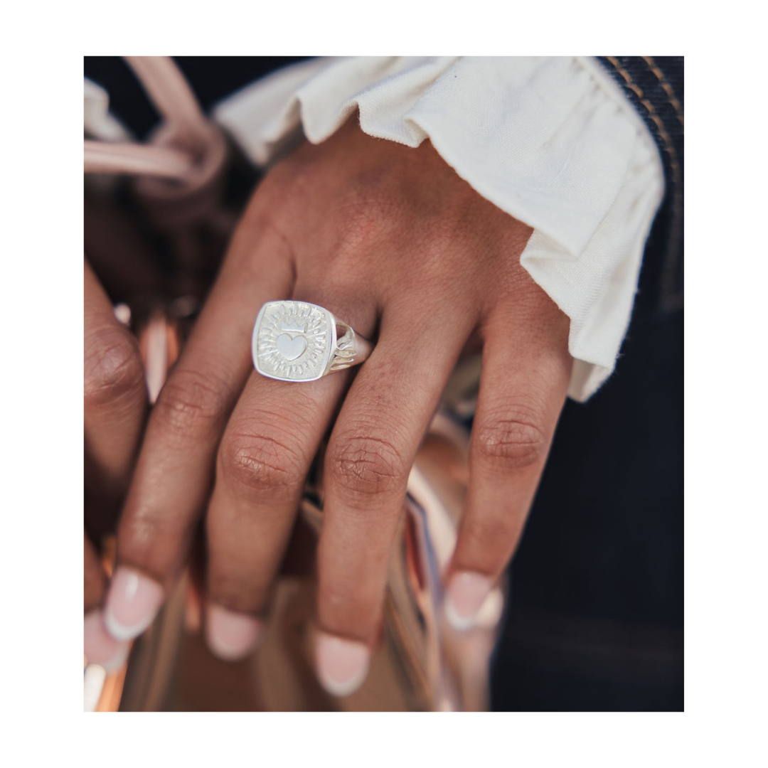 A close-up of a hand wearing a silver signet ring with an engraved circular face, styled with a white ruffled sleeve and a metallic bag in the background.