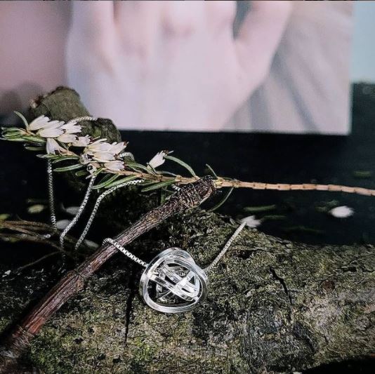 A Caroline Stokesberry-lee Knot Pendant Silver with a spherical, intricate design is displayed on a rustic tree branch adorned with small white flowers. The background is slightly blurred, showcasing part of a soft-focus photograph. The overall ambiance is earthy and artistic.