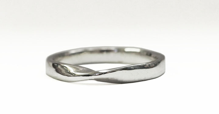 A close-up image of a simple silver twist shape ring. The ring has a smooth finish with a subtle twist design, introducing a gentle curve to its otherwise circular shape. The background is white, highlighting the reflective surface of this made-to-order Knot Wedding Ring by The Collective Dublin.