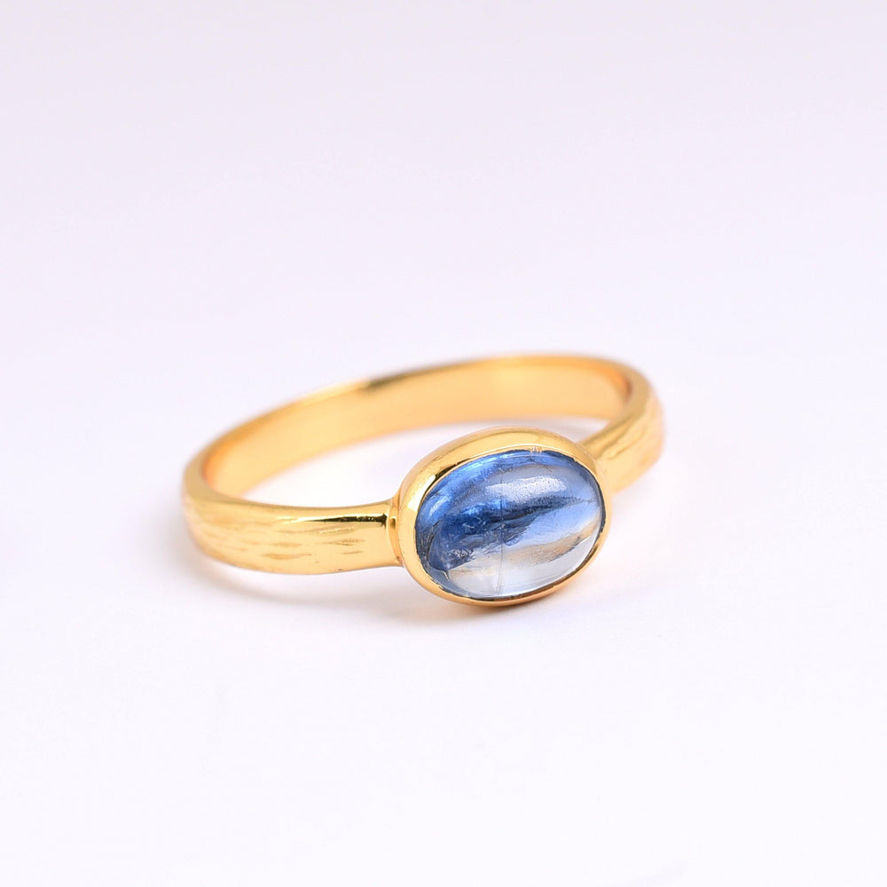 A gold plated sterling silver ring with a polished blue gemstone set in a simple, elegant band. The oval-shaped "Kyanite Gold Ring" by Watermelon Tropical has a glossy surface and subtle color variations, highlighted by the light. The ring is placed on a white background.