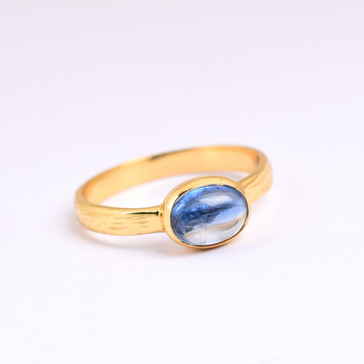 A gold plated sterling silver ring with a polished blue gemstone set in a simple, elegant band. The oval-shaped "Kyanite Gold Ring" by Watermelon Tropical has a glossy surface and subtle color variations, highlighted by the light. The ring is placed on a white background.