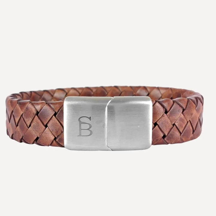 The Preston - Caramel from Steel and Barnett, a genuine leather, brown braided leather bracelet with a silver rectangular clasp engraved with a stylized "B" on a white background, adds an unmistakably masculine touch.