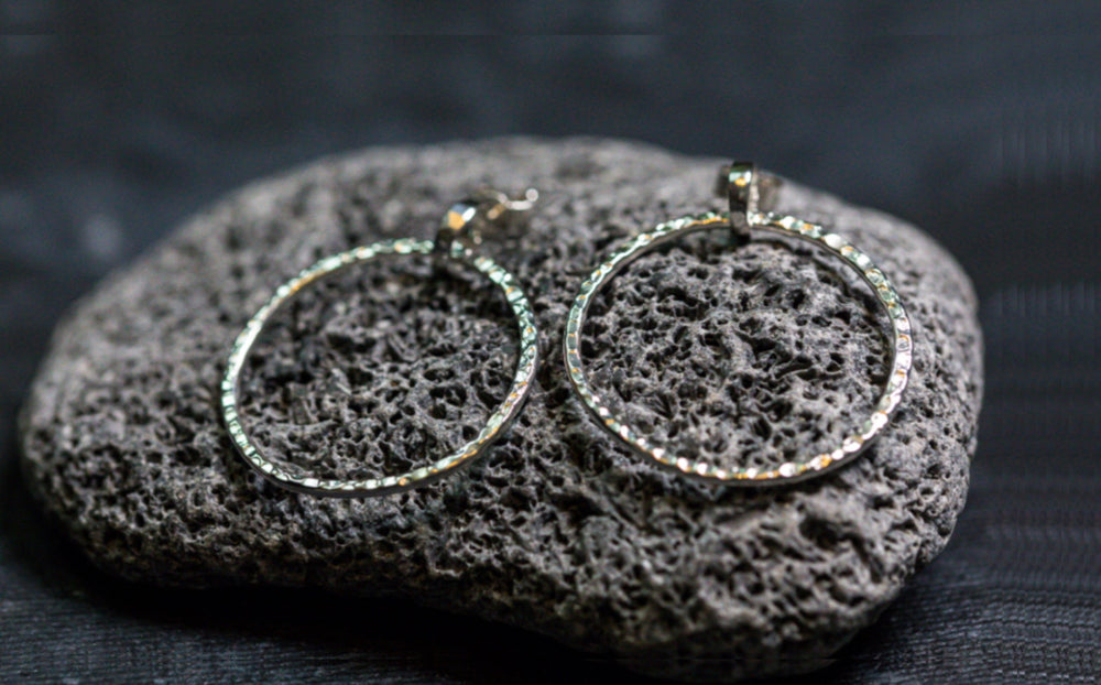 Close-up of a pair of Carran Hoops by Lynsey de Burca with a textured surface. The earrings are placed on a rough, dark gray volcanic rock, creating a striking contrast between the shiny metal and the matte stone. The background is out of focus, emphasizing the organic links in the design.