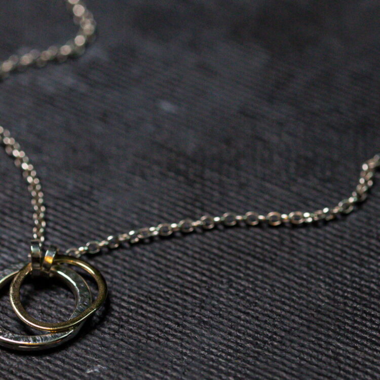 A delicate silver chain necklace with two intertwined rings as the pendant lies on a textured dark surface. One ring is silver and the other gold, symbolizing unity and connection. The close-up view captures the fine details and craftsmanship of the jewelry, perfectly complementing matching Carran earrings. This elegant piece is known as the **Carran Mix Pendant** by **Lynsey de Burca**.