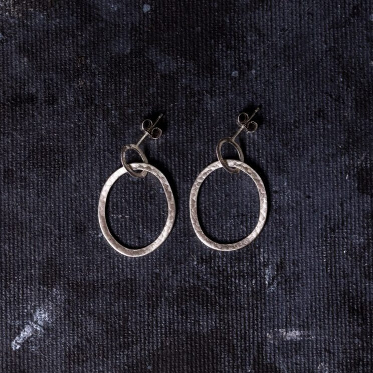 A pair of hand-forged Sterling silver earrings is displayed on a dark, textured background. The Carran Drops by Lynsey de Burca boast an irregular, hammered surface and are attached to stud posts. The overall look is minimalistic and modern.