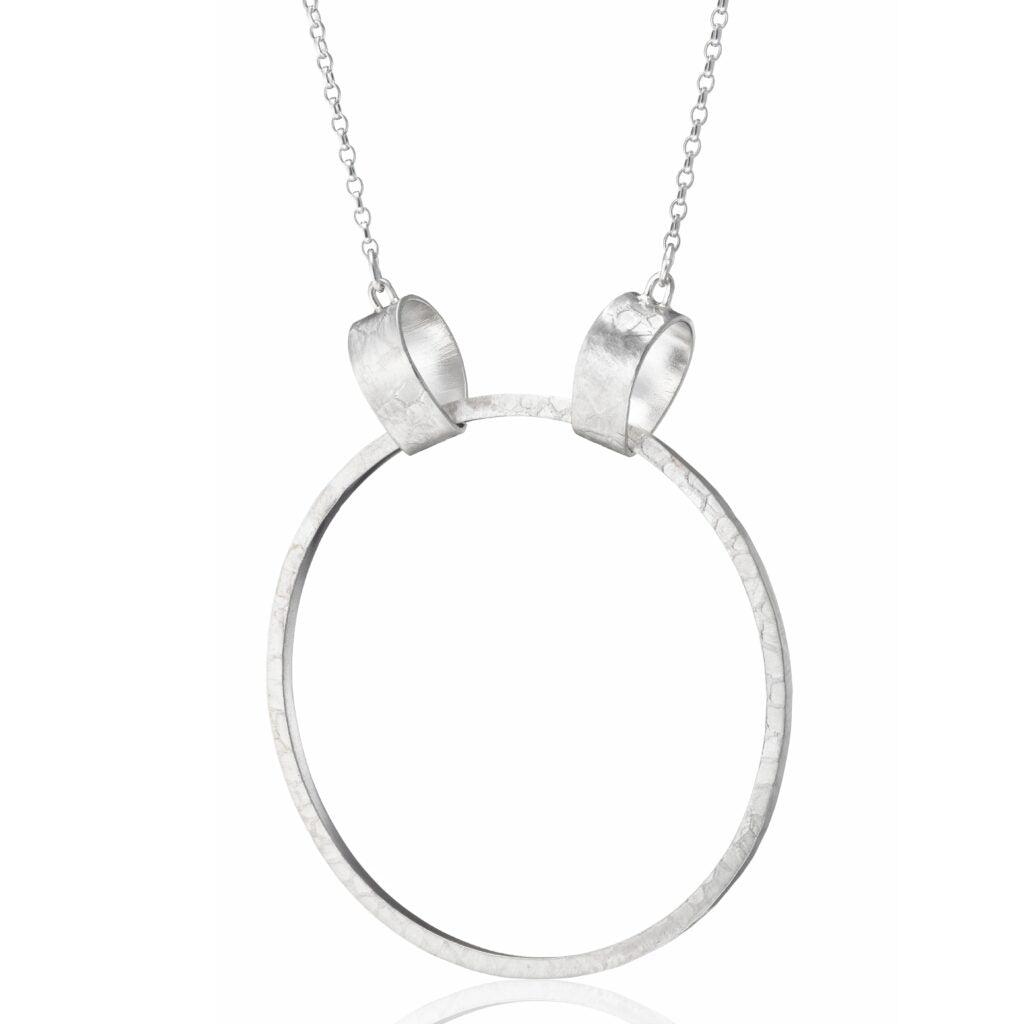 The Lynsey de Burca Talún Ancaire Neckpiece Silver is a minimalist sterling silver neckpiece featuring a single, large, textured circular pendant held by two small loops on a delicate chain. The pendant has a hammered finish, giving it a unique, handcrafted look. The thin and delicate chain adds an elegant touch.