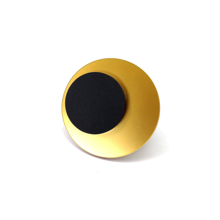 A round, minimalist object with a sleek gold outer ring and a smaller, black circular center. The design is simple, with smooth surfaces and a modern aesthetic. Hand-polished to perfection, the Lina Ring by Alex + Svet exudes elegance against the white background.