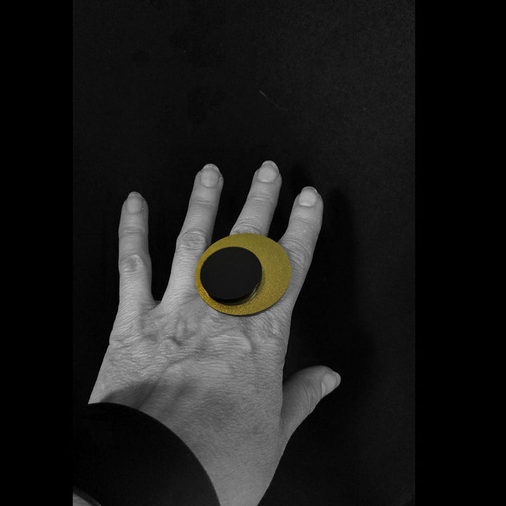 A hand, with a black and gold circular Lina Ring from Alex + Svet on its middle finger, is positioned against a black background. The hand-polished ring has two concentric circles—the smaller one black—superimposed on a larger gold circle. The image is in monochrome except for the meticulously crafted ring.