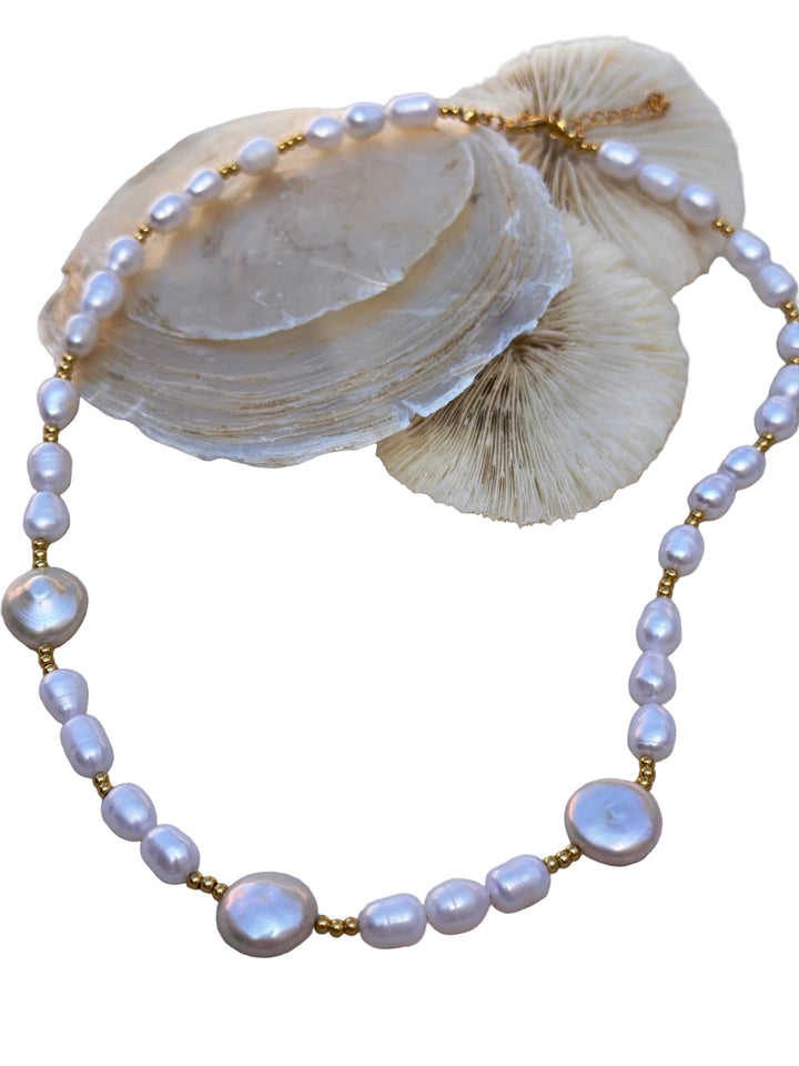 The La Perla Necklace by Kybalion Jewellery features irregular white freshwater pearls and gold beads with a gold-plated clasp, handmade in Dublin. It's shown draped on light seashells and coral against a white background.