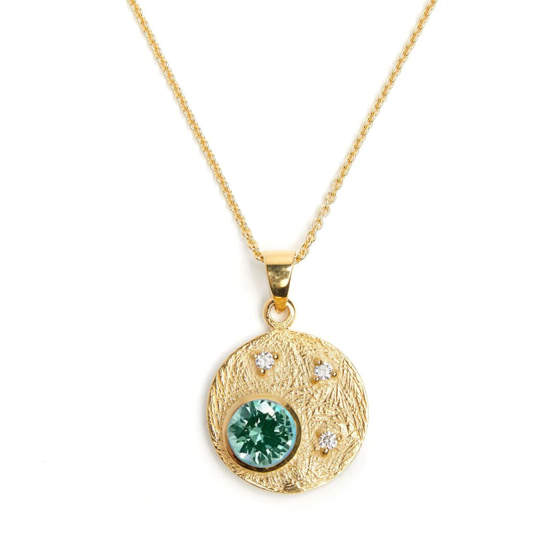 The Gallardo & Blaine Designs Celeste Necklace in Gold features a round pendant with a central green gemstone and three smaller cubic zirconia on a textured surface, elegantly paired with a simple gold link chain against a pristine white background.