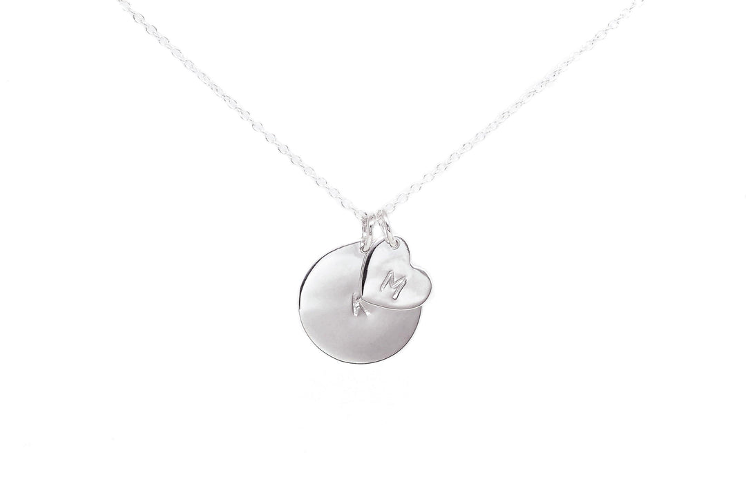 A Personalised Charms Personalised Large Disc Charm in Silver featuring two pendants on a delicate chain. One pendant is a smooth, round disc, and the other is a small heart with the letter "M" hand stamped on it. The image has a white background, emphasizing the simplicity and elegance of this personalized gift.